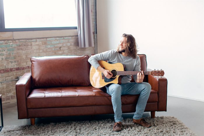 Man with long hair sitting on brown leather couch holding a guitar while strumming.