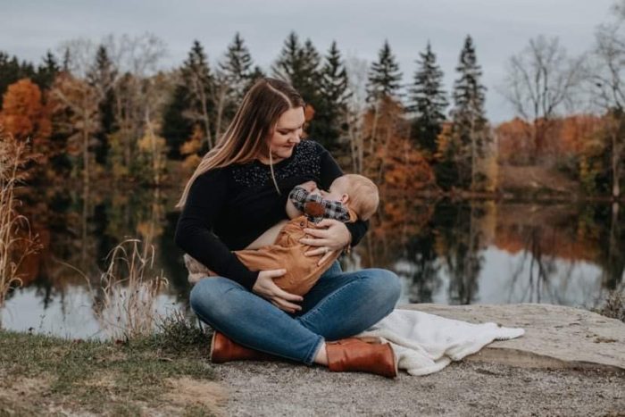 Woman with long hair in jeans seated cross-legged and holding baby to breastfeed
